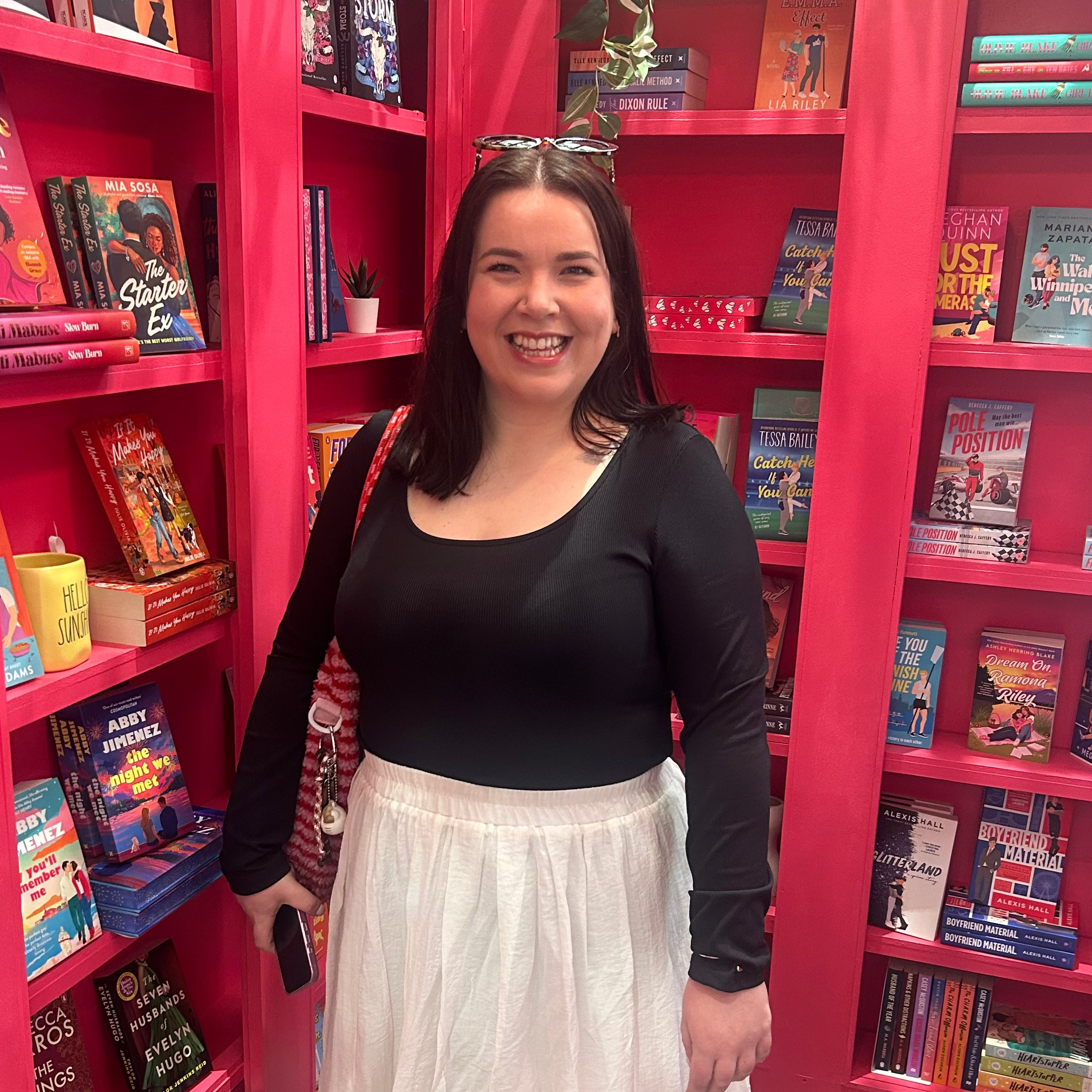 A woman smiling in a bookstore, wearing a black long-sleeve top and a long white skirt, standing in front of bright pink bookshelves filled with books.