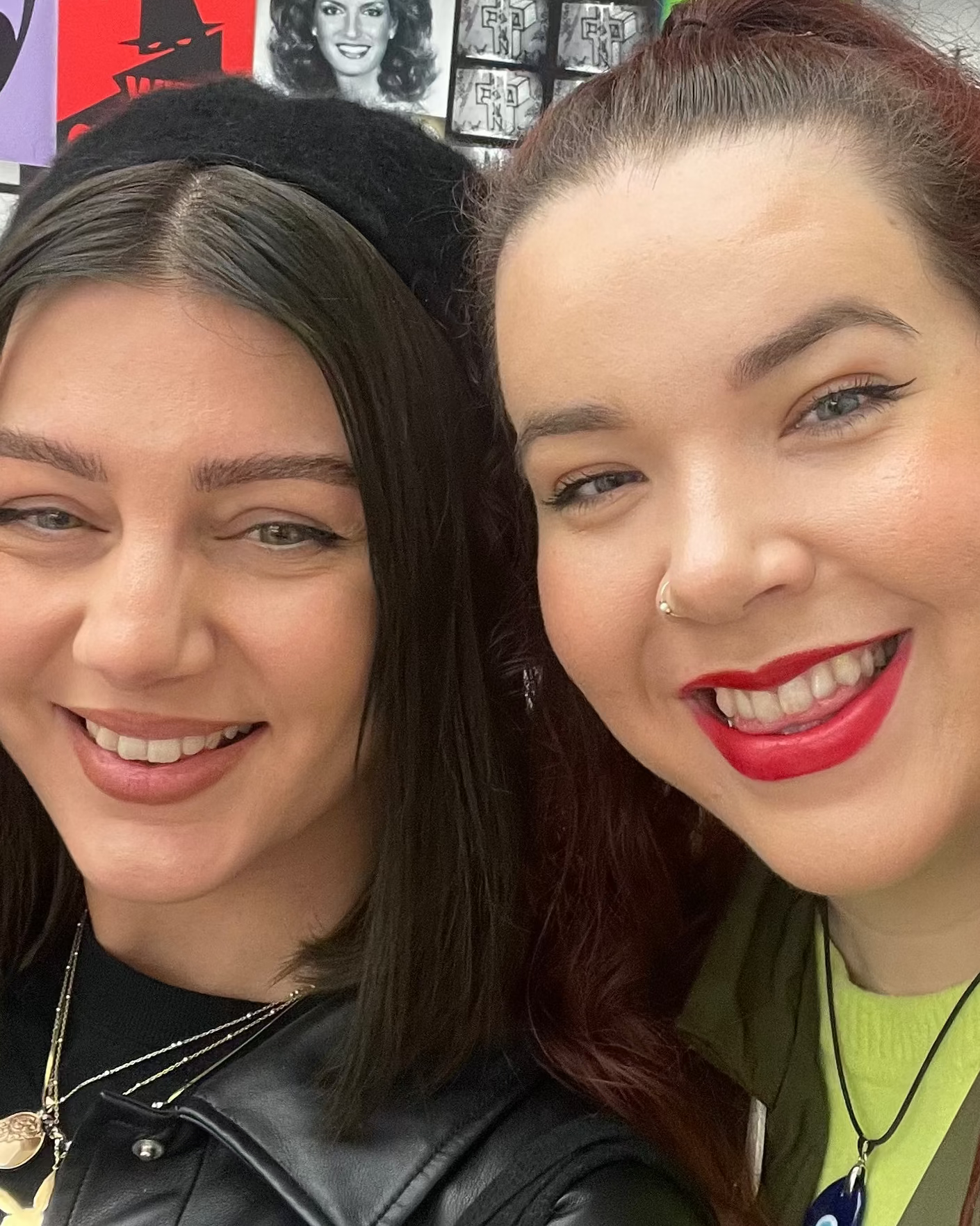 Two women smiling for a selfie in front of a colorful collage wall. The woman on the left has dark hair and wears a black leather jacket, while the woman on the right has reddish-brown hair and wears an olive green jacket over a neon yellow top.