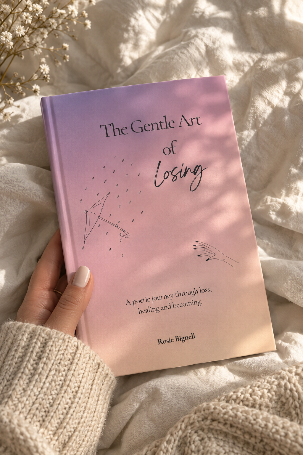 A hand in a cream knit sweater holds a book titled "The Gentle Art of Losing" by Rosie Bignell. The book cover is a soft pink and lavender gradient with minimalist line drawings, set against a cozy background of white textured bedding with soft sunlight and shadows.