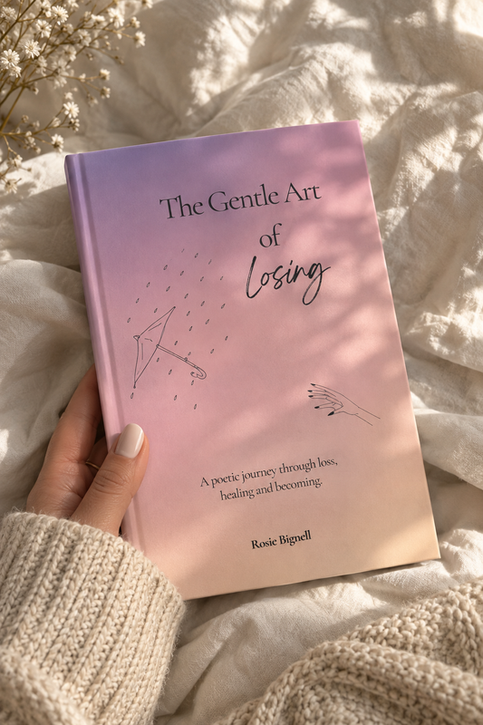 A hand in a cream knit sweater holds a book titled "The Gentle Art of Losing" by Rosie Bignell. The book cover is a soft pink and lavender gradient with minimalist line drawings, set against a cozy background of white textured bedding with soft sunlight and shadows.